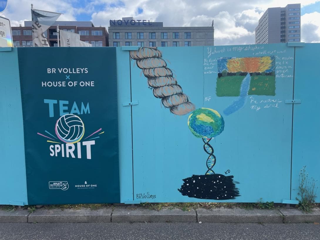 Das Banner "House of One x BR Volleys" mit einem Volleyball zwischen den Worten Teamspirit hängt neben dem fertigen Kunstwerk der Profis der BR Volleys. Eine Erde die aus drei verschiedenen Quellen - hier die Bilder der Spieler - gehalten wird. 