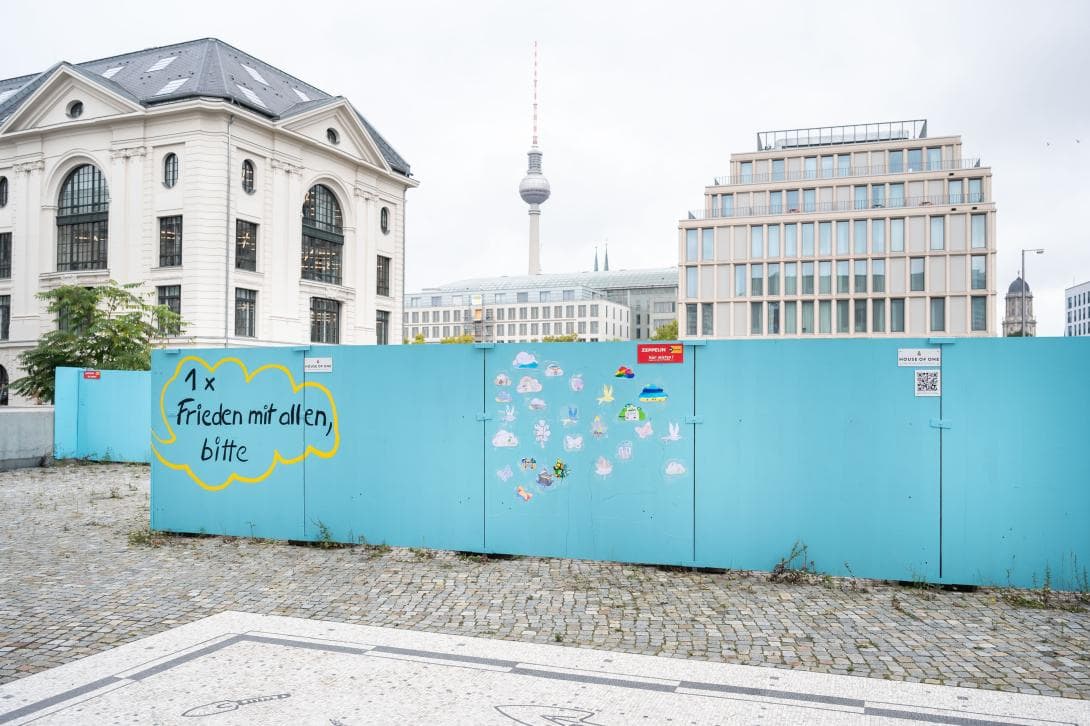 Blick auf den Bauzaun des House of One, auf dem die Nachricht "1x Frieden mit allen, bitte" zu lesen und die Friedensbilder der Schüler zu sehen sind. Im Hintergrund ist der Fernsehturm auf dem Alexanderplatz zu sehen.