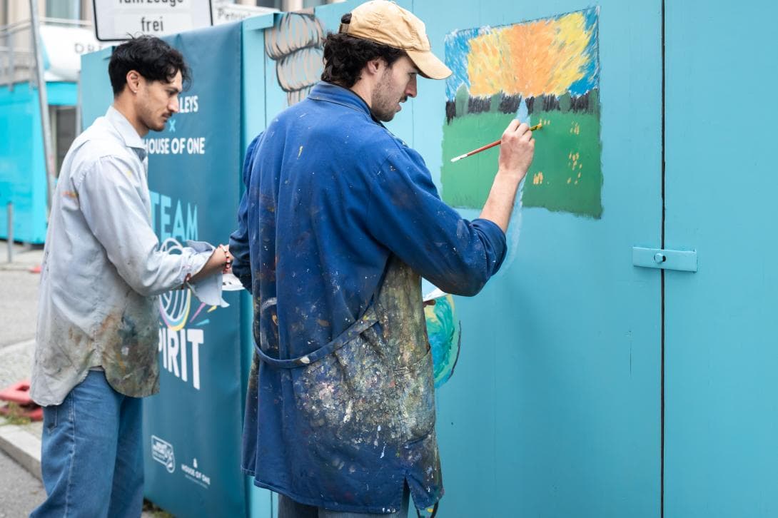 Nehmiah Mote und Nolan Flexan, Profi-Spieler der BR Volleys, malen ihre Botschaften auf den Bauzaun des House of One. 